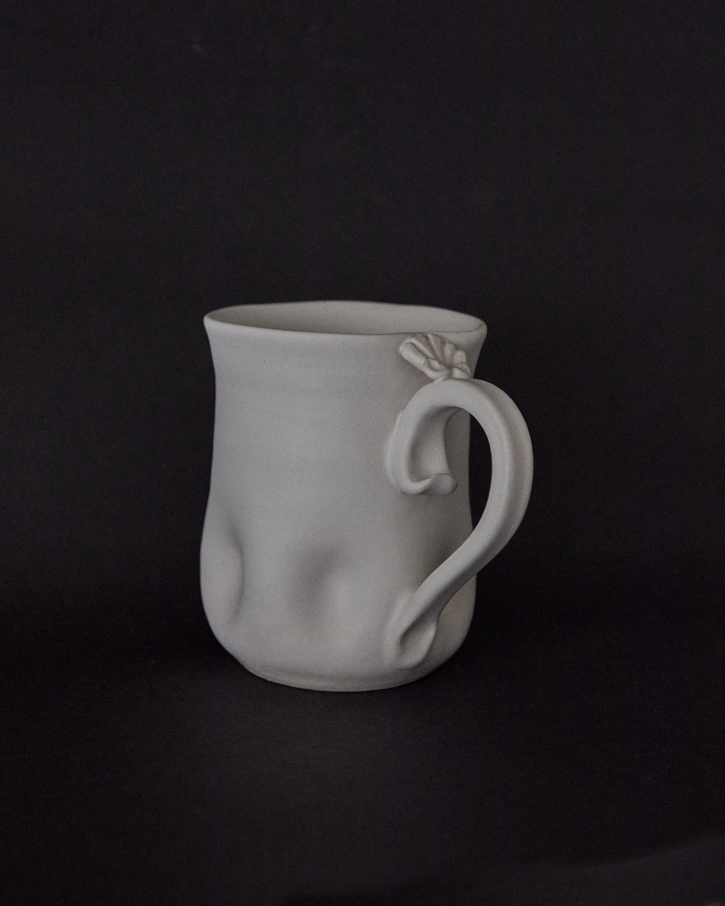 A white ceramic mug with a sculptural, organic handle and subtle textured dimples on its body, set against a solid black background.