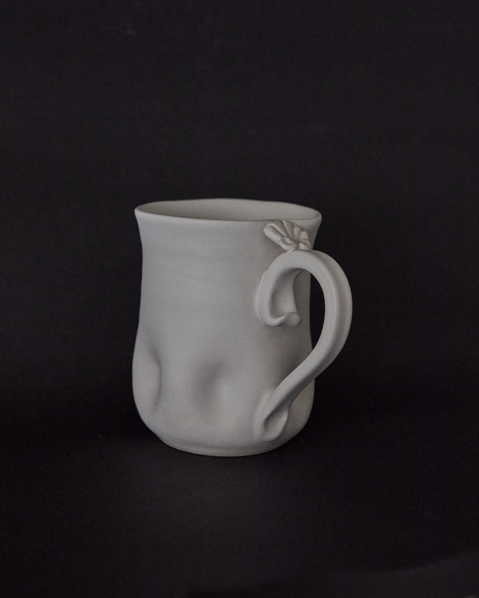 A white ceramic mug with a sculptural, organic handle and subtle textured dimples on its body, set against a solid black background.